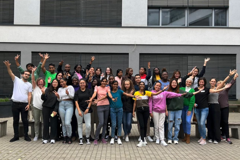 A large group of international language students stands together in front of a language school in Heidelberg, smiling and laughing at the camera.
