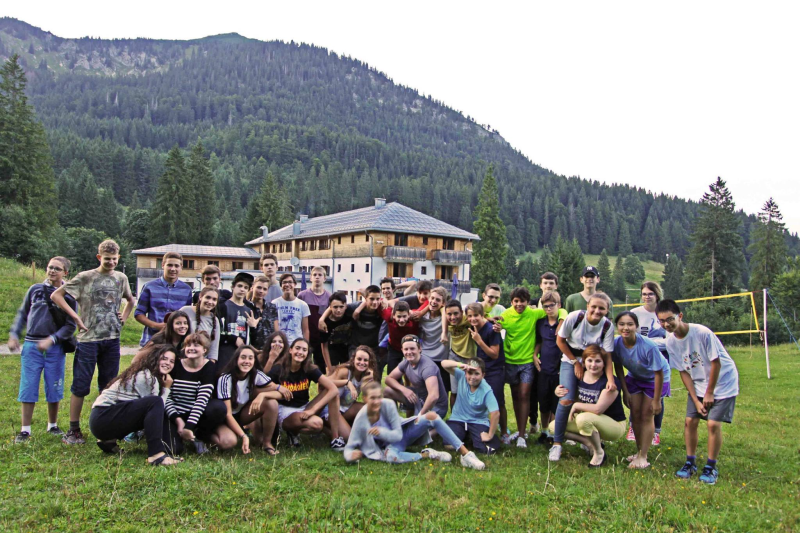 Jugendliche Teilnehmer des Sommercamps München Spitzingsee stehen gemeinsam auf einer Wiese in den Bergen nahe München und lachen in die Kamera.